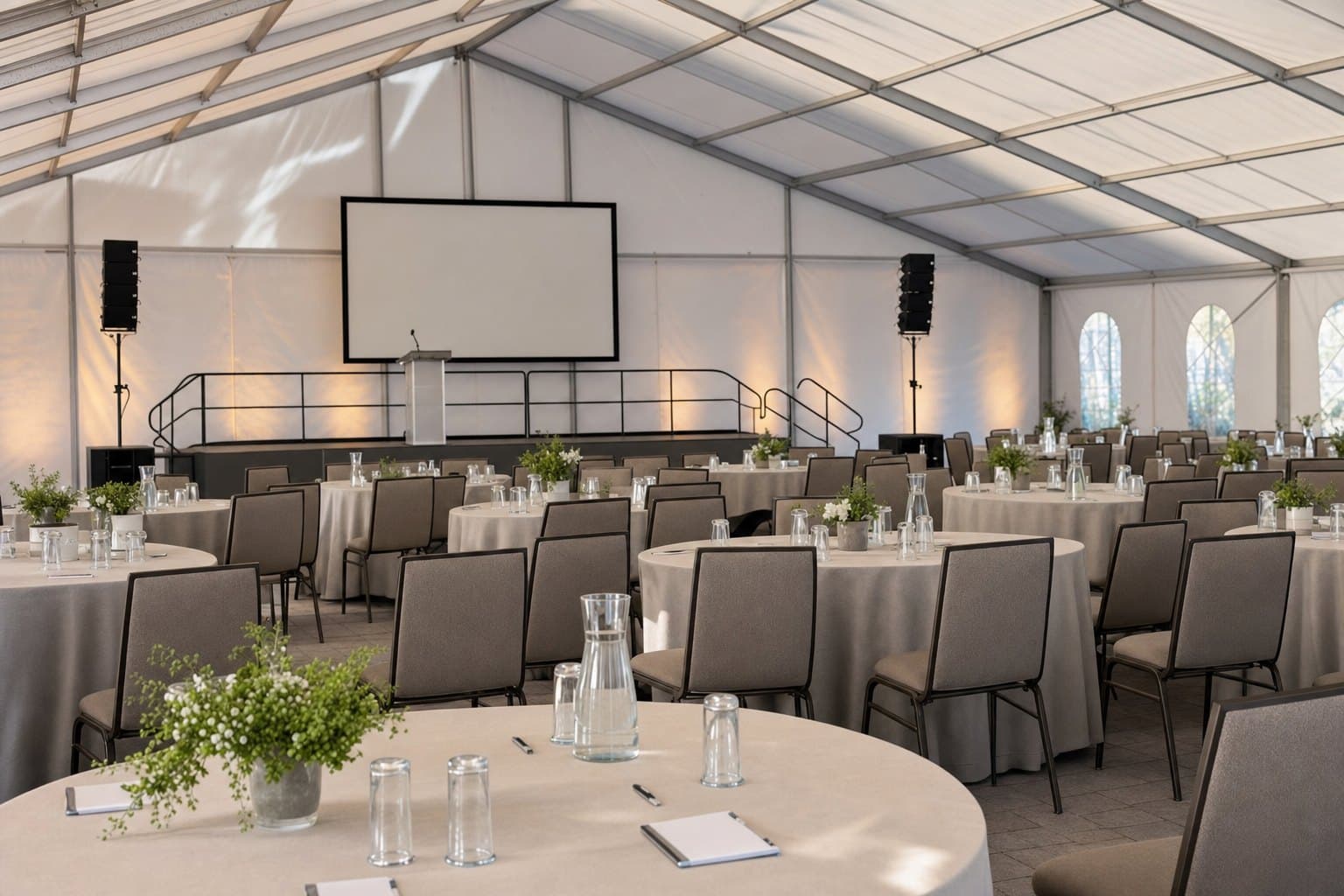 Corporate event setup under a clear-span tent with rows of banquet tables facing a stage and presentation screen by Creative Events & Rentals